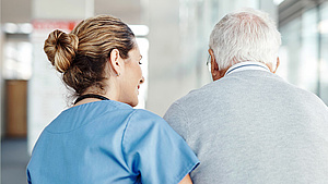 Eine Krankenschwester begleitet einen älteren Mann beim Gehen durch einen Flur.