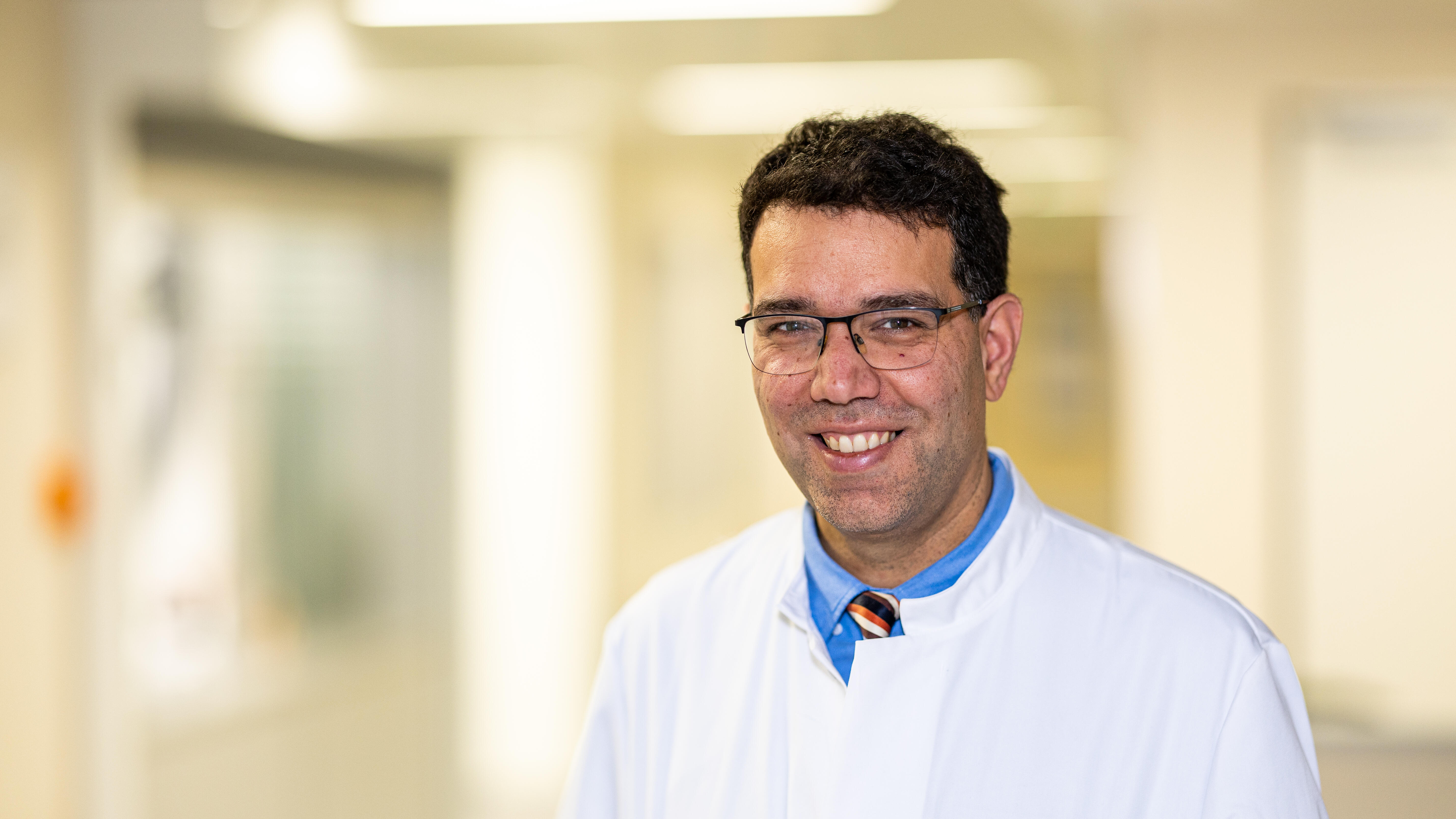 Ein braunhaariger Mann mittleren Alters mit Brille lächelt im Aztkittel freundlich in die Kamera. Der Krankanhausflur im Hintergrund ist hell aber verschwommen.