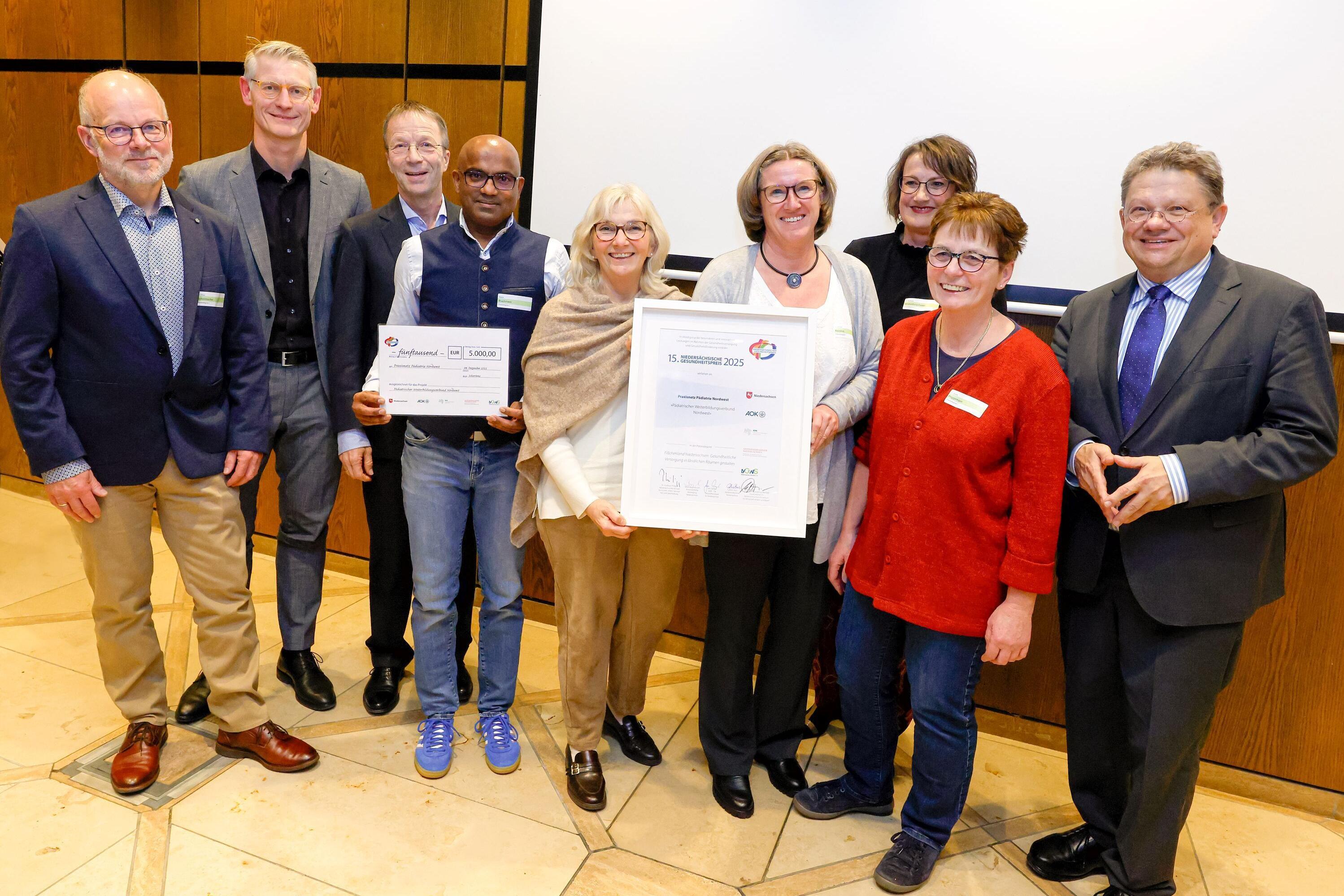 Neun Personen stehen im Halbkreis, vier Frauen in der Mitte, rechts davon Gesundheitsminister Philippi mit dreieckigen Handhaltung Angela Merkels, vier Männer stehen links von der Frauengruppe. Alle lächeln, eine Frau und ein Mann in der Mitte halten jeweils eine Urkunde hoch. 