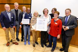 Neun Personen stehen im Halbkreis, vier Frauen in der Mitte, rechts davon Gesundheitsminister Philippi mit dreieckigen Handhaltung Angela Merkels, vier Männer stehen links von der Frauengruppe. Alle lächeln, eine Frau und ein Mann in der Mitte halten jeweils eine Urkunde hoch. 