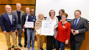 Neun Personen stehen im Halbkreis, vier Frauen in der Mitte, rechts davon Gesundheitsminister Philippi mit dreieckigen Handhaltung Angela Merkels, vier Männer stehen links von der Frauengruppe. Alle lächeln, eine Frau und ein Mann in der Mitte halten jeweils eine Urkunde hoch. 