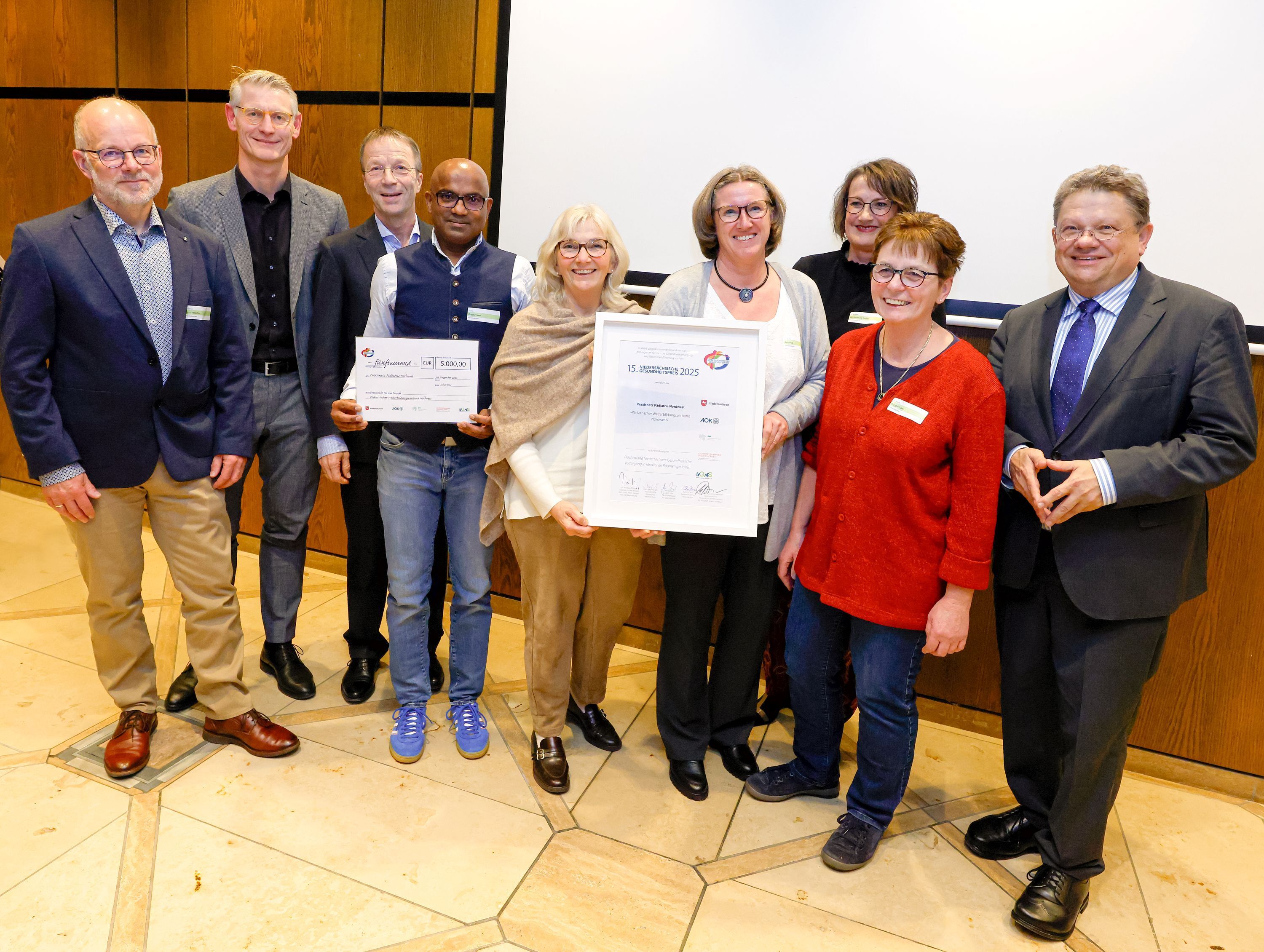 Neun Personen stehen im Halbkreis, vier Frauen in der Mitte, rechts davon Gesundheitsminister Philippi mit dreieckigen Handhaltung Angela Merkels, vier Männer stehen links von der Frauengruppe. Alle lächeln, eine Frau und ein Mann in der Mitte halten jeweils eine Urkunde hoch. 